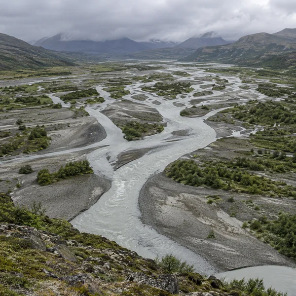 A braided river splits into many pale channels across a broad gravel floodplain dotted with low green scrub, with dark hills and cloud in the distance.