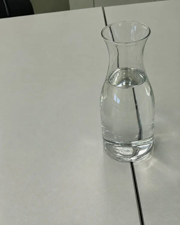 A clear glass carafe partly filled with water stands over the dark seam between pale grey tabletop panels, with the seam visibly refracted through the glass…