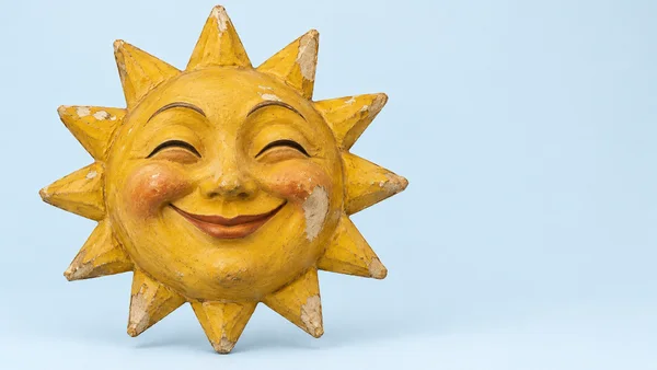 A worn yellow papier-mâché sun ornament with a smiling face and triangular rays sits against a pale blue background, leaving open space on the right.