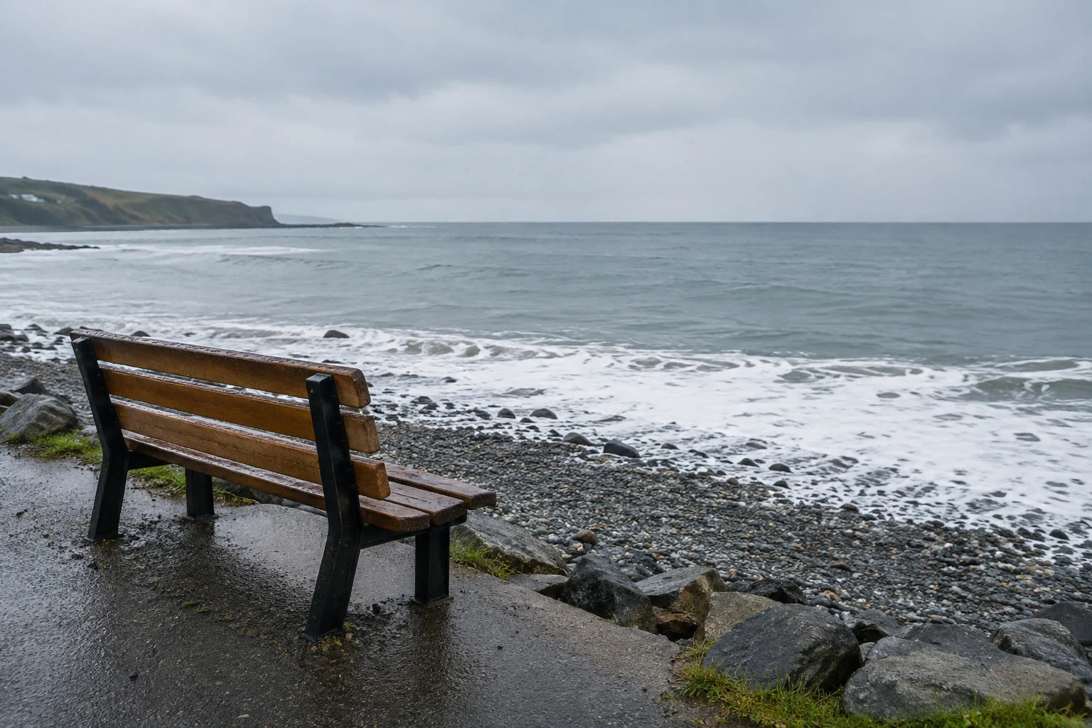 A weathered brown wooden bench stands on a wet coastal path above dark pebbles, facing a grey sea where white foam breaks along the shore beneath an overcast…