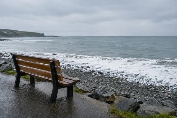 A weathered brown wooden bench stands on a wet coastal path above dark pebbles, facing a grey sea where white foam breaks along the shore beneath an overcast…