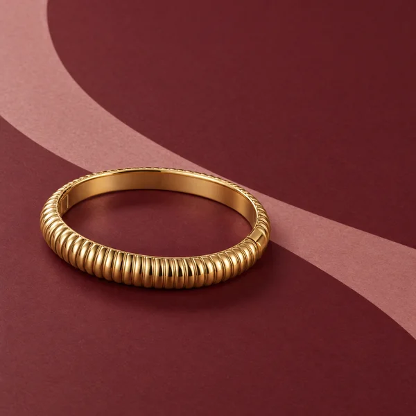 A ribbed gold bangle rests on a wine-coloured surface, with a broad blush-coloured curve sweeping diagonally behind it.