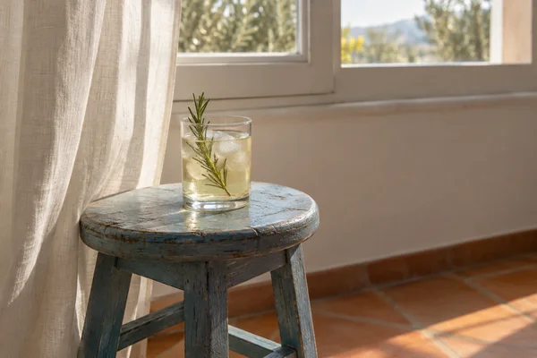 A clear tumbler of pale golden tonic with ice and a rosemary sprig sits on a weathered blue wooden stool beside an off-white curtain and sunlit window…