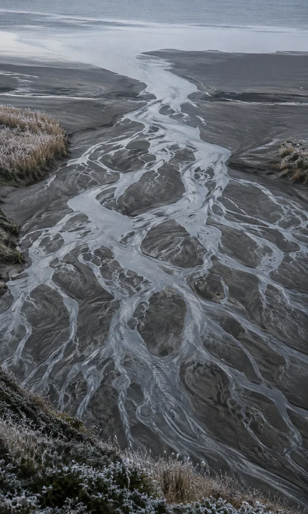 A high view looks down a dark sandy river delta, where a broad pale channel branches into many thin silver waterways.