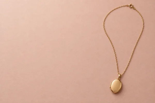 A fine gold necklace lies in an elongated loop on blush-pink textured paper, with an oval locket near the lower right and a clasp at the top; the left half is…