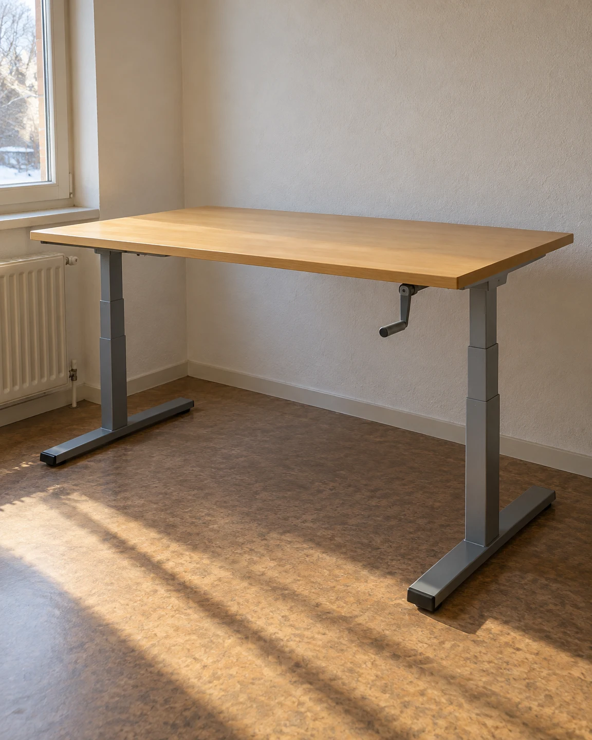 An empty birch-top height-adjustable desk with grey telescoping legs and a small hand crank stands in a sunlit room beside a window, above a mottled brown…