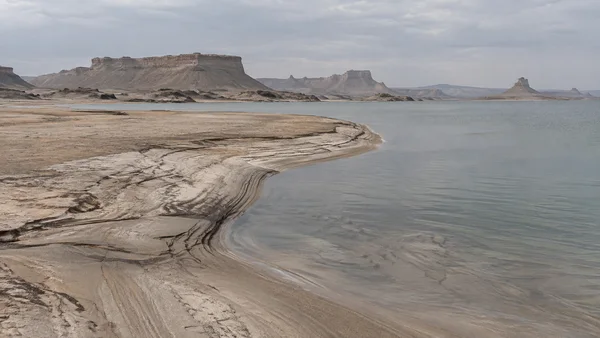A pale, layered sand spit curves from the left foreground into calm grey water, with eroded flat-topped mesas and sculpted ridges across the distant horizon…