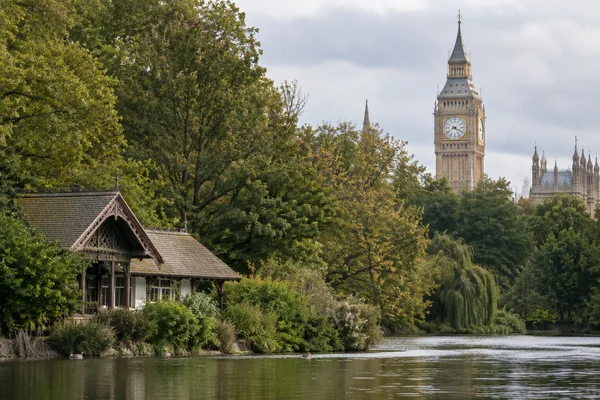 A timber chalet-style cottage sits among dense lakeside planting beside calm water, with Elizabeth Tower and part of the Palace of Westminster rising beyond…