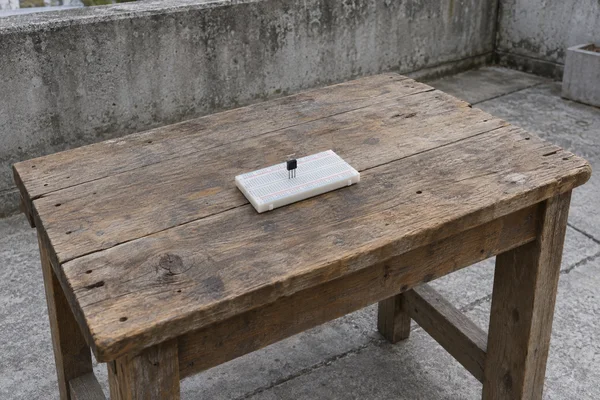 A small black transistor stands upright on a white solderless breadboard near the center of a weathered wooden workbench, with a concrete wall and paving…