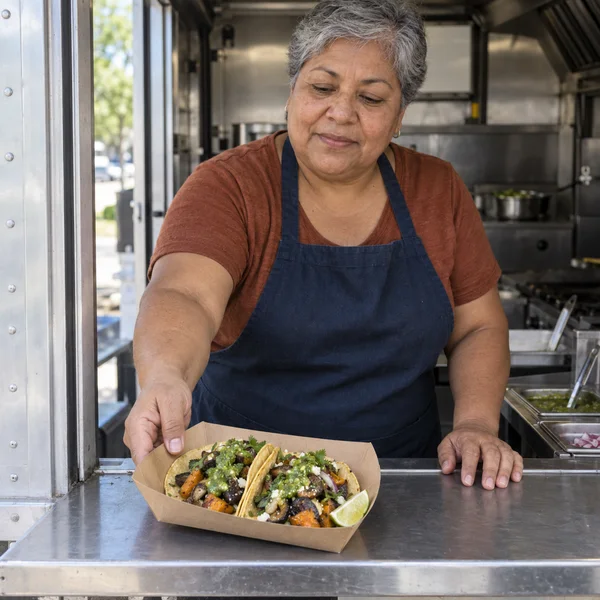 A gray-haired woman in a navy apron and russet shirt serves a kraft-paper tray holding two vegetable tacos with green salsa through a stainless-steel…