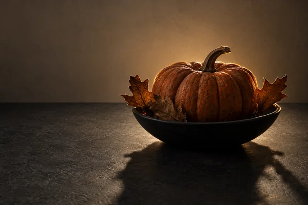 A large ribbed burnt-orange pumpkin with a curved stem sits in a shallow black bowl among dry oak leaves on a dark tabletop, with a warm stone-coloured…