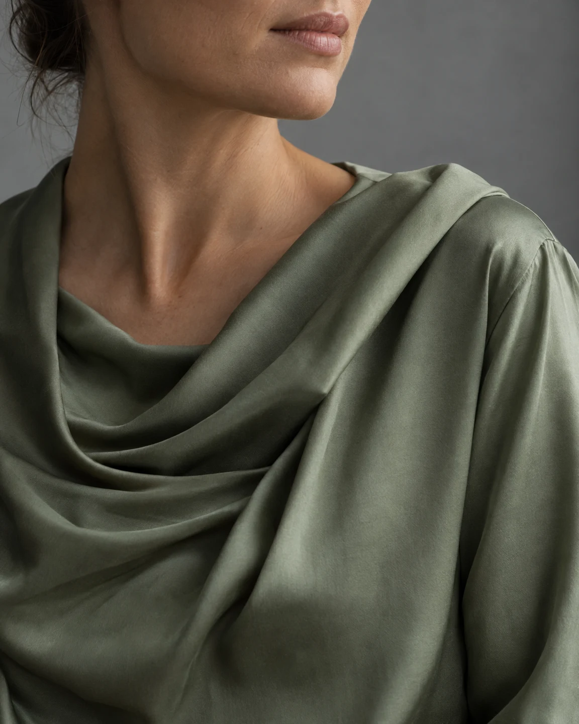 Tight studio portrait of an adult with relaxed lips and a turned profile, wearing a muted sage-green blouse with a deep, asymmetrical cowl neckline and broad…