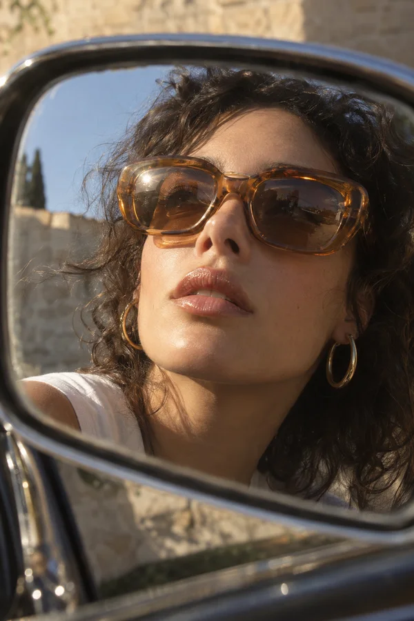 Close-up reflection of a woman with tousled dark curls, oversized amber sunglasses, gold hoop earrings, and a white top in a curved chrome car side mirror…