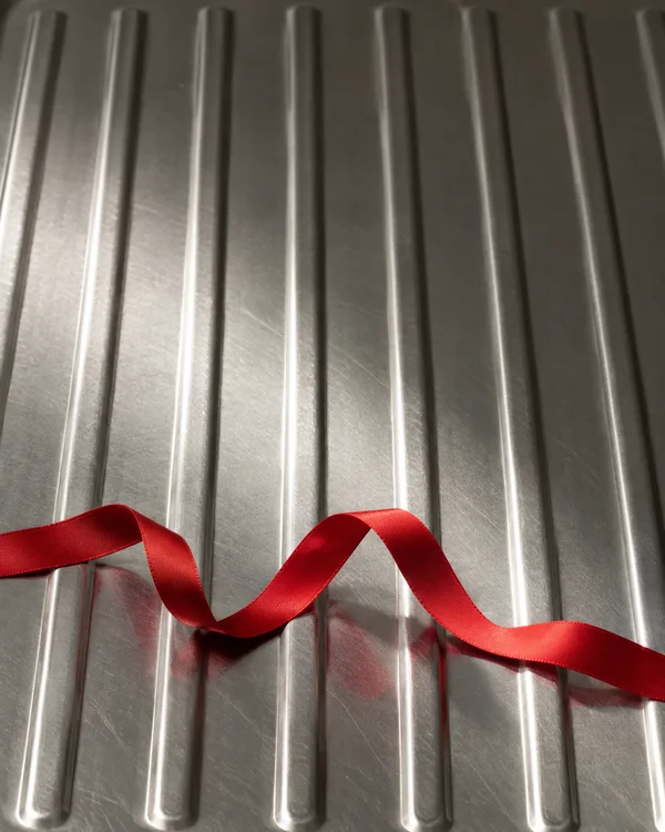 A loose red satin ribbon snakes across the lower third of a vertically ridged stainless-steel draining board, with reflections visible in the metal and open…