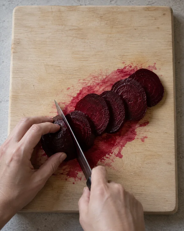 Overhead view of hands slicing a cooked beetroot on a pale wooden chopping board, with dark burgundy rounds arranged in a curved row over a broad smear of red…