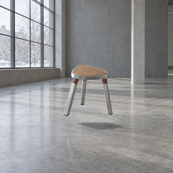 A three-legged stool with a rounded triangular cork seat and silver metal legs floats above a concrete floor in a spacious grey studio.