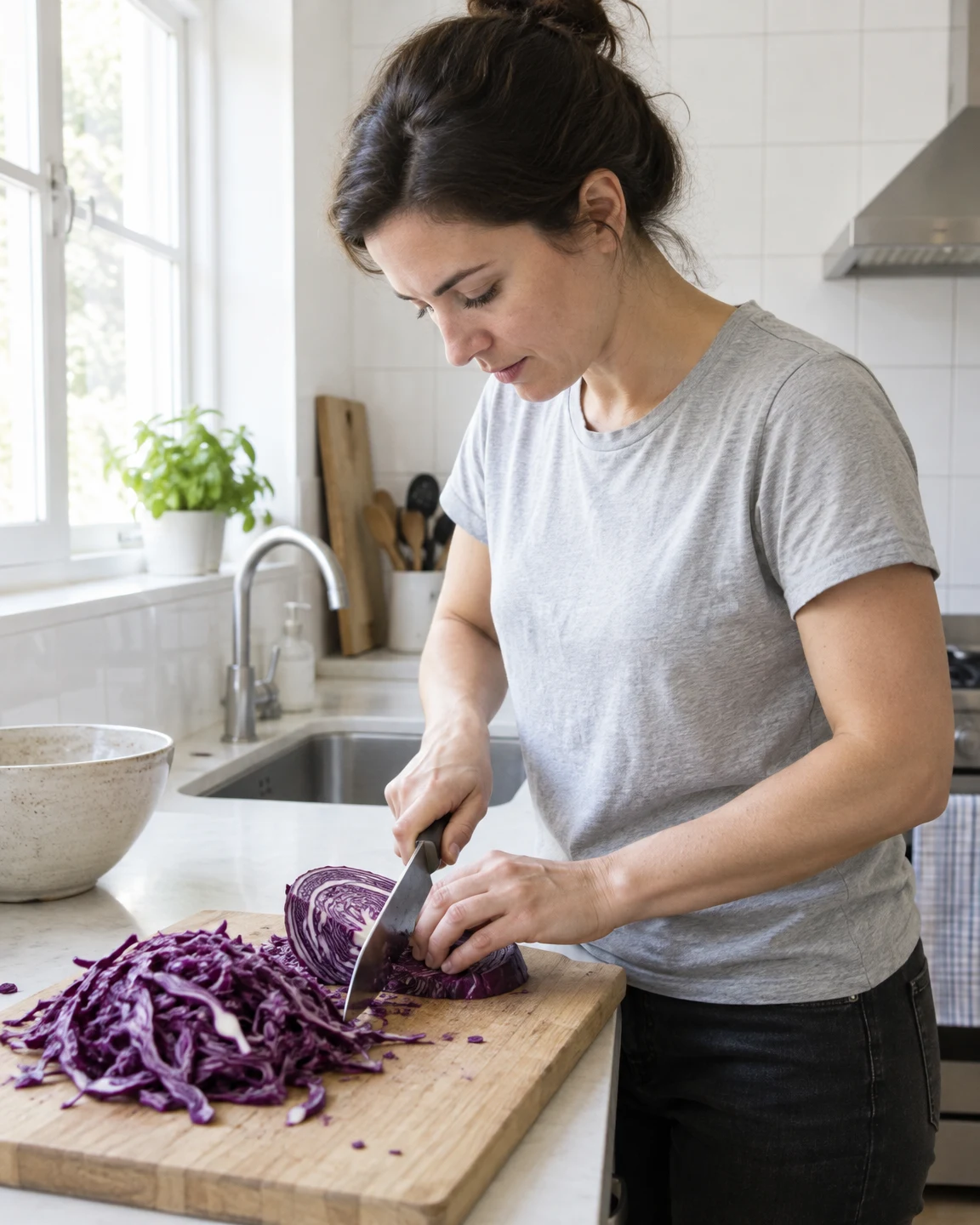 A woman in a pale-grey T-shirt leans over a kitchen counter, chopping a halved red cabbage on a wooden cutting board beside a large pile of purple shreds.