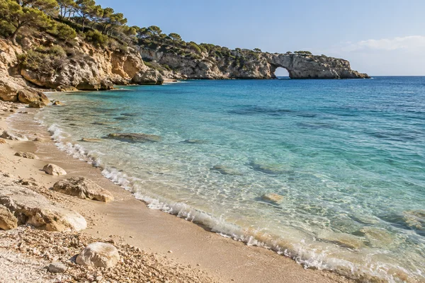A quiet limestone cove with pale pebbly sand curving beside clear aquamarine shallows and blue water.