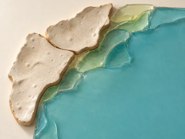Overhead assemblage of irregular ivory ceramic fragments and translucent cyan and pale yellow glass pieces arranged as a curving lakeshore on a warm ivory…