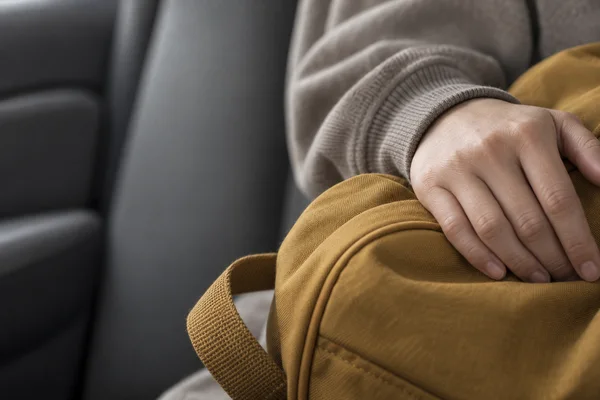 A cropped hand in a warm-gray sleeve rests on a muted-ochre fabric backpack inside a car, with a loose webbing strap and blurred gray upholstery filling the…