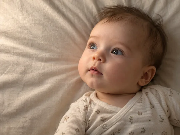 Close overhead three-quarter portrait of an awake infant lying on pale oatmeal-colored cotton, gazing upward with a relaxed mouth and wearing a light…