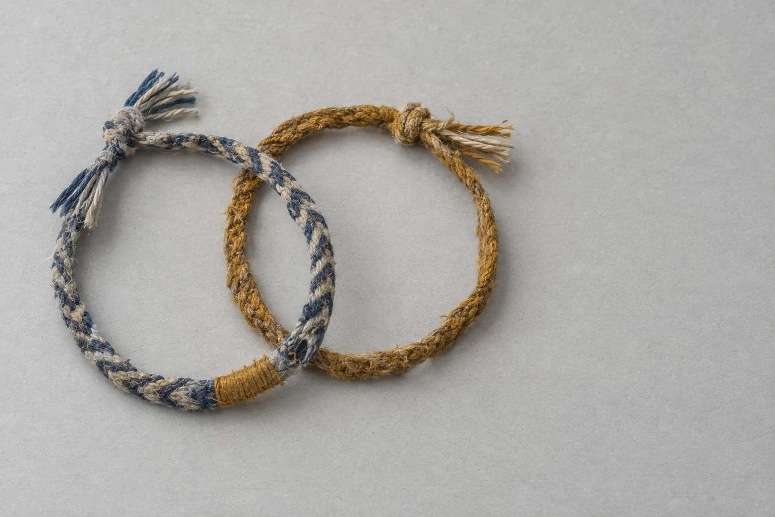 Two worn cotton friendship bracelets lie in overlapping open loops on a pale grey surface.