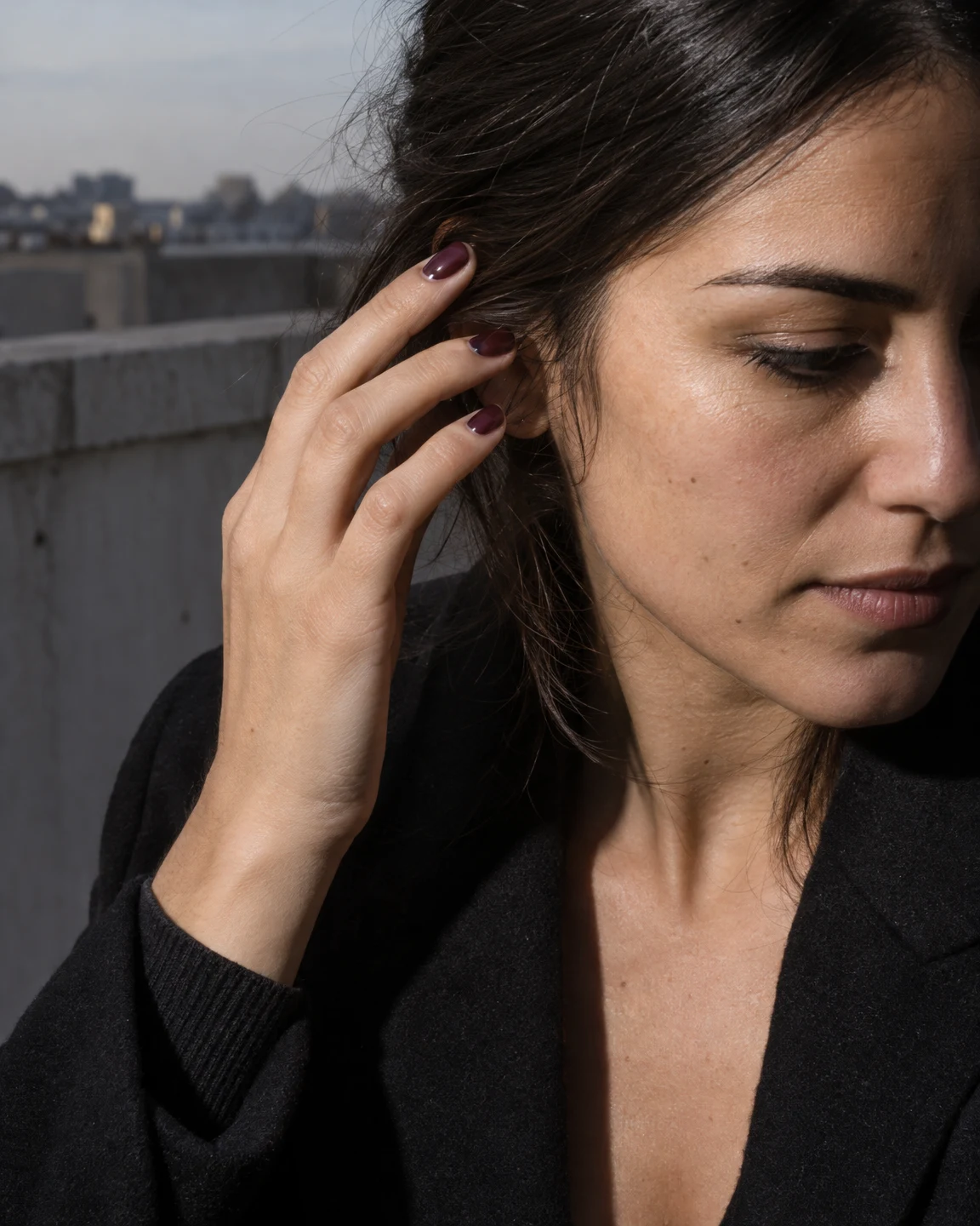 A close vertical portrait of a dark-haired adult looking downward while lifting a hand to tuck hair behind one ear.