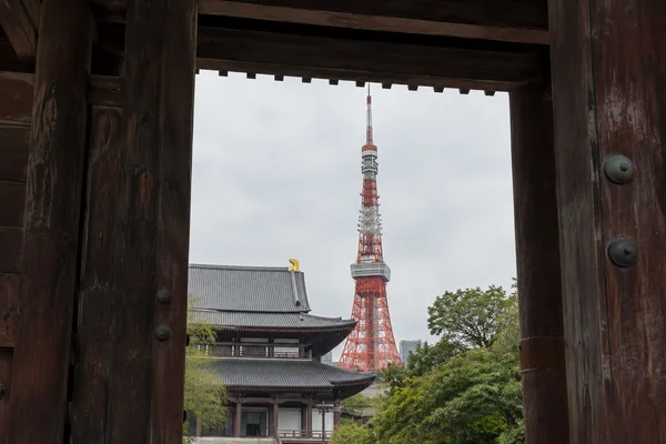 Tokyo Tower rises behind traditional temple roofs, framed by the dark wooden posts and overhead beam of a large gate beneath a pale gray sky.