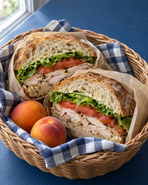 Two cut halves of a roast chicken, tomato, and lettuce sourdough sandwich sit in a wicker basket lined with a navy-and-white checked napkin and parchment, with…