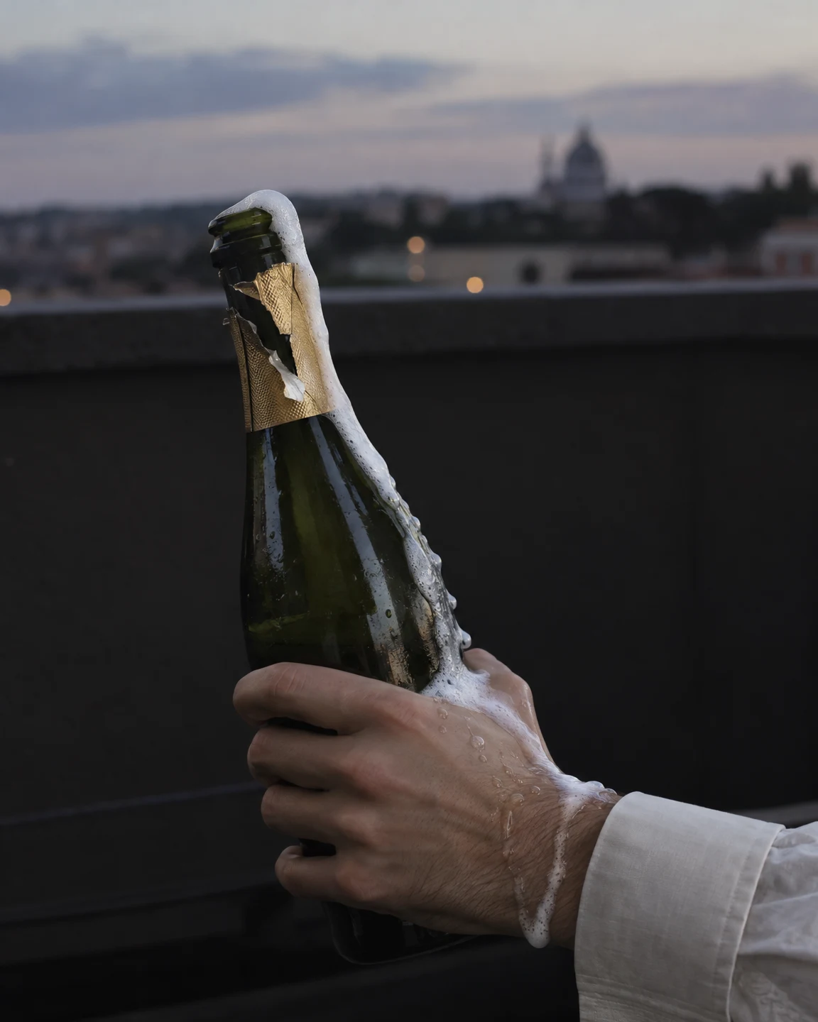 A hand grips a dark green sparkling-wine bottle as white foam spills over its neck, down the glass and across the wrist beside an ivory cuff; a softly blurred…