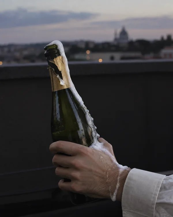 A hand grips a dark green sparkling-wine bottle as white foam spills over its neck, down the glass and across the wrist beside an ivory cuff; a softly blurred…