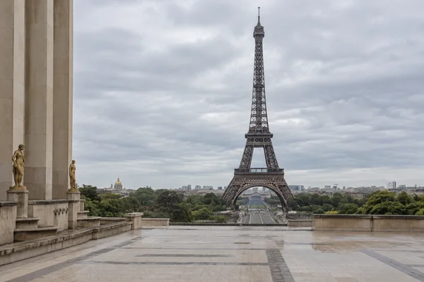 The Eiffel Tower rises beyond the Trocadéro gardens, viewed across a broad stone terrace bordered by a pale limestone façade and statues beneath an overcast…