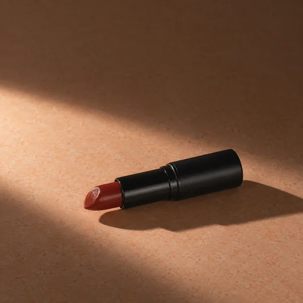 A brick-red lipstick in a plain black tube lies diagonally across a peach tabletop, lit by a broad diagonal band of light and casting a long shadow.