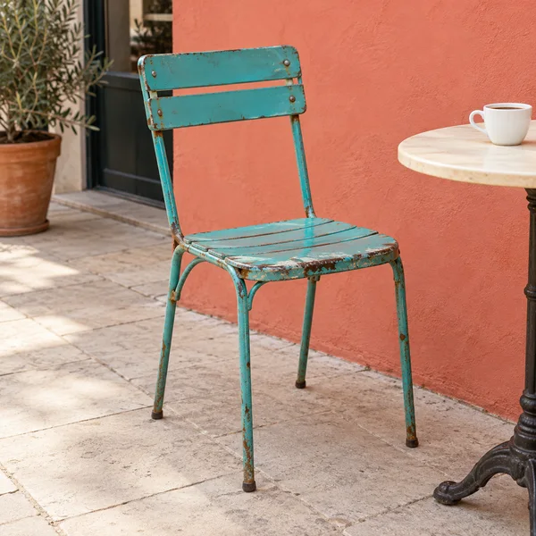 A weathered turquoise metal chair with chipped paint and rust stands on pale tiled pavement against a coral wall.