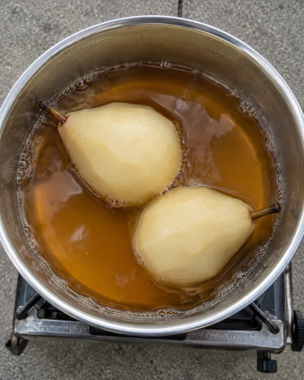 An overhead view of two peeled pears with stems simmering in amber liquid in a stainless-steel pot on a portable burner over gray concrete.