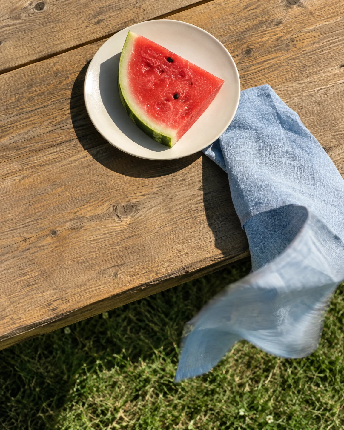 An overhead view of a triangular watermelon wedge on an off-white plate beside a pale blue linen napkin, on a sunlit wooden table with grass visible beyond its…