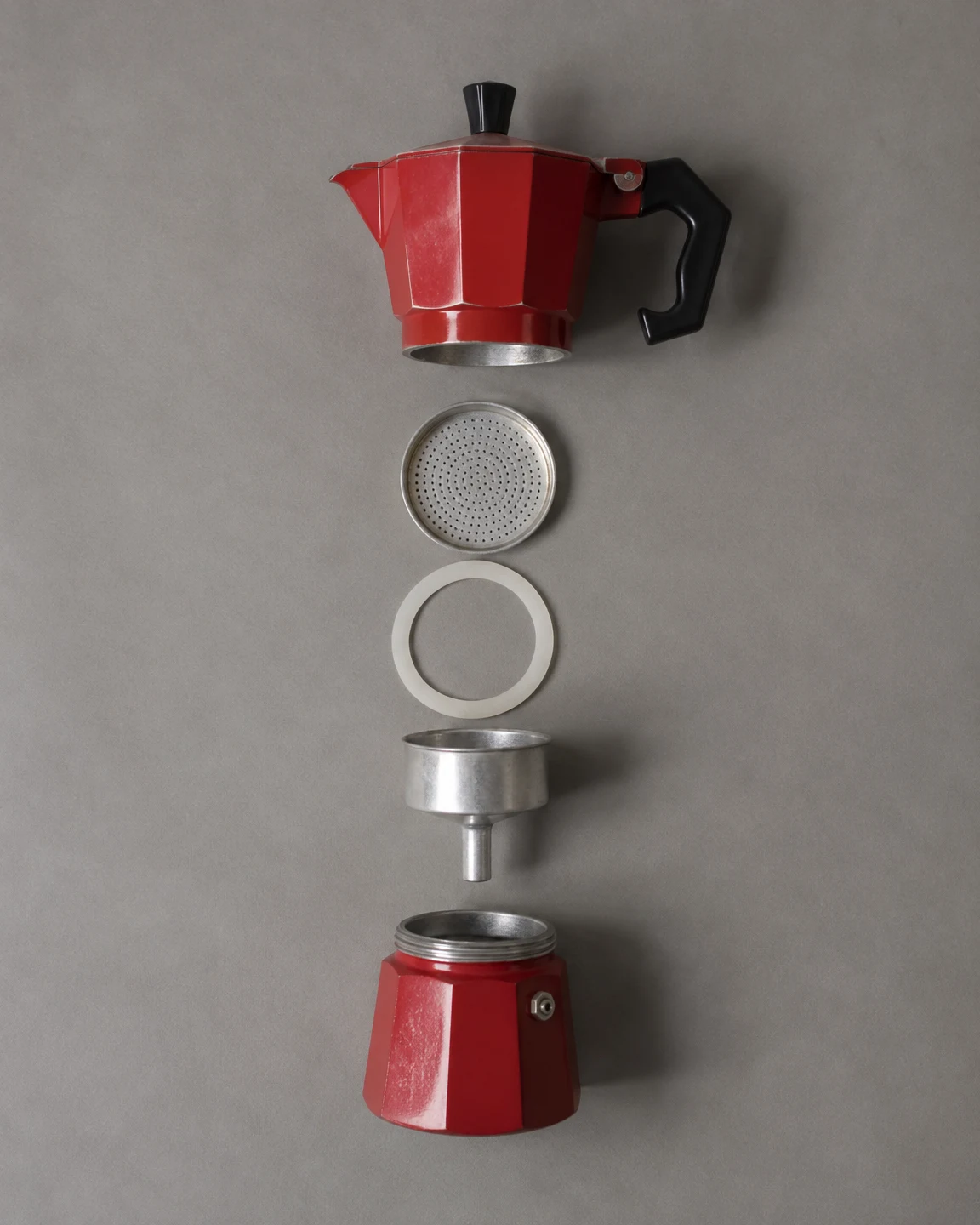 An overhead flat lay of a red moka pot dismantled into its upper chamber, perforated filter plate, pale gasket, metal funnel basket and lower boiler, arranged…