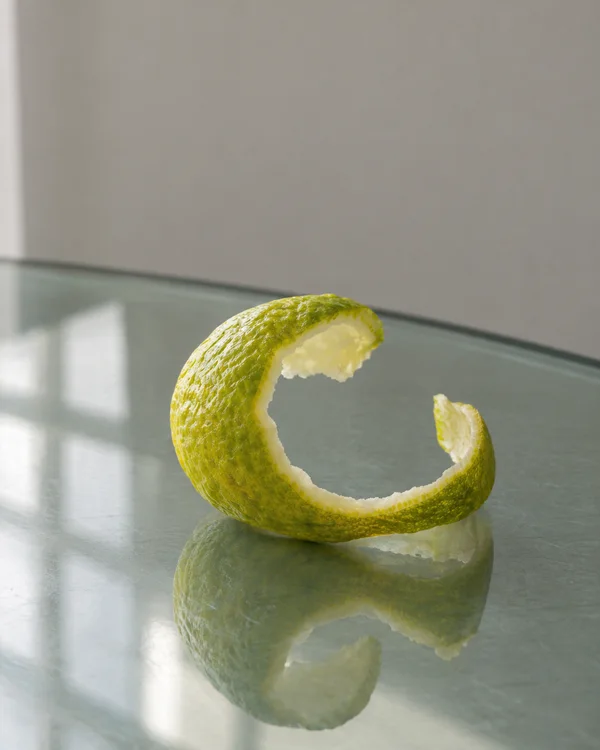 A yellow-green citrus peel curls into an open ring on a reflective glass tabletop, with creamy pith along its cut edge and a soft reflection below.