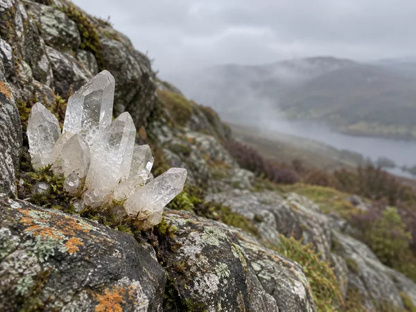 A translucent quartz crystal cluster rises from a lichen- and moss-speckled grey rock ledge, with one point angled outward; a softly blurred misty hillside…