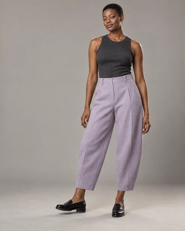 A full-length woman with closely cropped hair stands against a warm-gray studio backdrop, wearing a charcoal sleeveless top, pale-lilac ankle-length trousers…