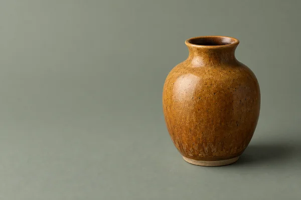 A speckled amber ceramic vase with a narrow, flared neck and pale unglazed foot sits on a muted sage-gray surface, with open space to its left and a soft…
