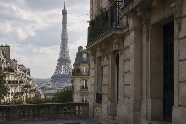 The Eiffel Tower rises beyond Parisian rooftops, framed by a weathered stone townhouse with wrought-iron balconies on the right and a stone balustrade across…