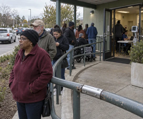 A line of people waits along a curved ramp outside a clinic entrance as cars pass nearby.
