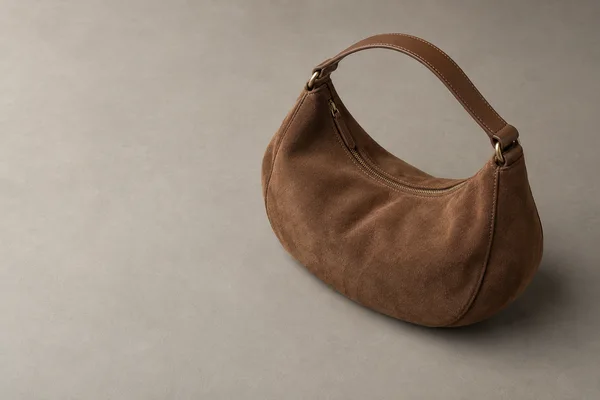 A chestnut-brown suede crescent handbag with a stitched leather handle and brass zipper sits on a warm-grey tabletop, leaving broad empty space to the left.