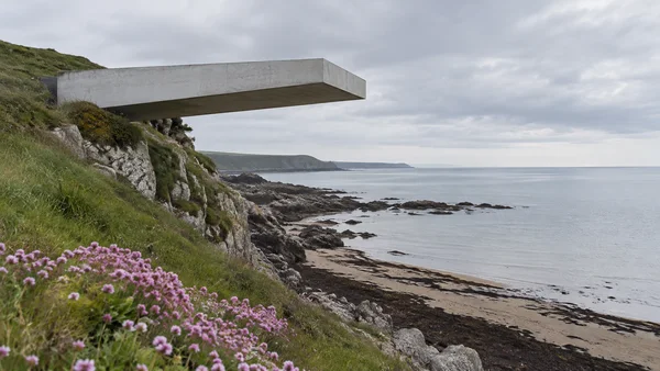 A pale concrete platform projects from a grassy, rocky coastal headland above a dark, kelp-streaked shore and calm grey sea.