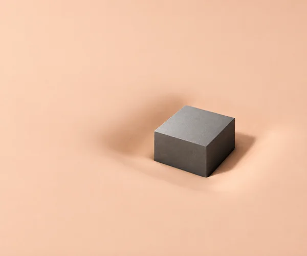 A graphite-grey cuboid rests slightly right of centre in a shallow depression on a matte pale-apricot surface, with a short shadow falling to the right and…