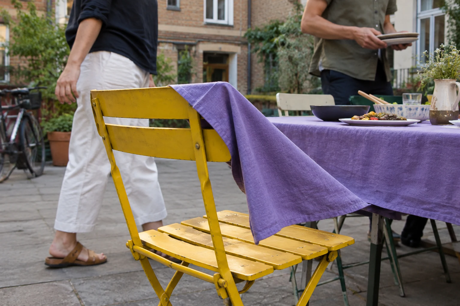 A yellow folding chair fills the foreground of a paved courtyard, with a violet tablecloth draped from the shared dining table over its backrest.