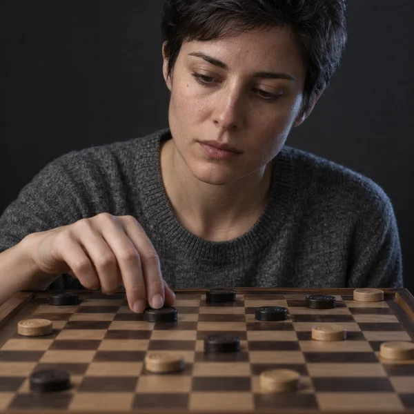 A short-haired adult in a charcoal knit top looks down while moving a dark round piece on a wooden checkered board, against a dark studio background.