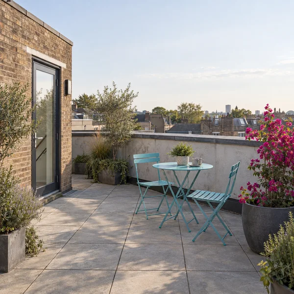 A cyan round bistro table and two matching folding chairs sit on a paved rooftop terrace beside a large charcoal planter filled with bright pink flowers.