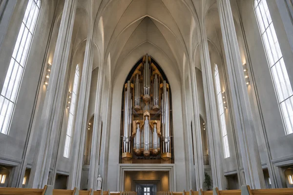 A centered view down a large church nave toward a tall pipe organ above a doorway, framed by pale ribbed concrete vaults and tall side windows; wooden pews…
